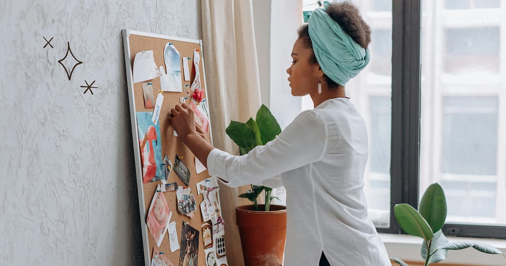 A woman creating a financial vision board at home, pinning inspirational images and goals onto a corkboard. The room is bright and cozy, with natural light coming through the window and plants adding a touch of greenery