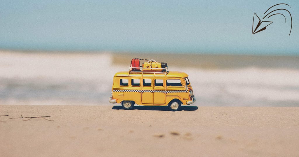 A miniature yellow van with luggage on top sits on a sandy beach, with a blurred ocean backdrop under a bright blue sky, symbolizing travel, exploration, and fresh starts. Summer vacation from school, which to budget for a new start.