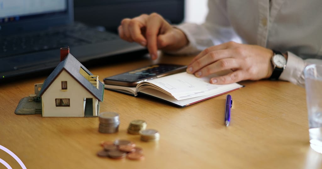 Person calculating finances at a desk with coins, a notebook, and a small house model, representing fair loan practices and financial planning.