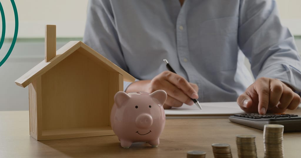 A person calculating finances at a desk with a piggy bank, coins, and a wooden house model, illustrating how payroll linked loans work to simplify repayments through salary deductions.