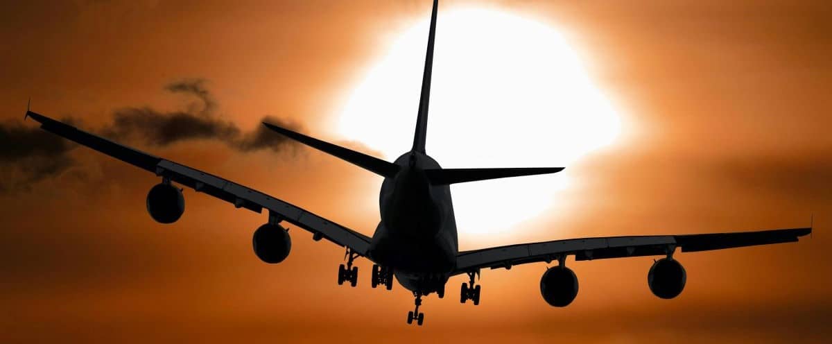 A silhouette of an airplane in flight against a dramatic orange sky, with the sun glowing brightly in the background, partially obscured by clouds that will make people Save Money