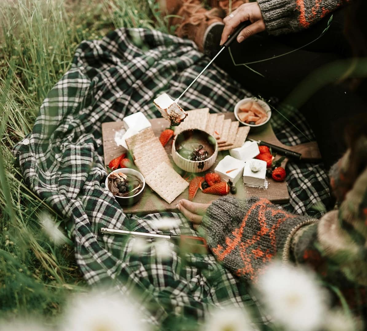 A rustic picnic setup with a checkered blanket and a charcuterie board featuring crackers, strawberries, marshmallows, and melted chocolate. A person in a cozy sweater roasts a marshmallow over a flame.