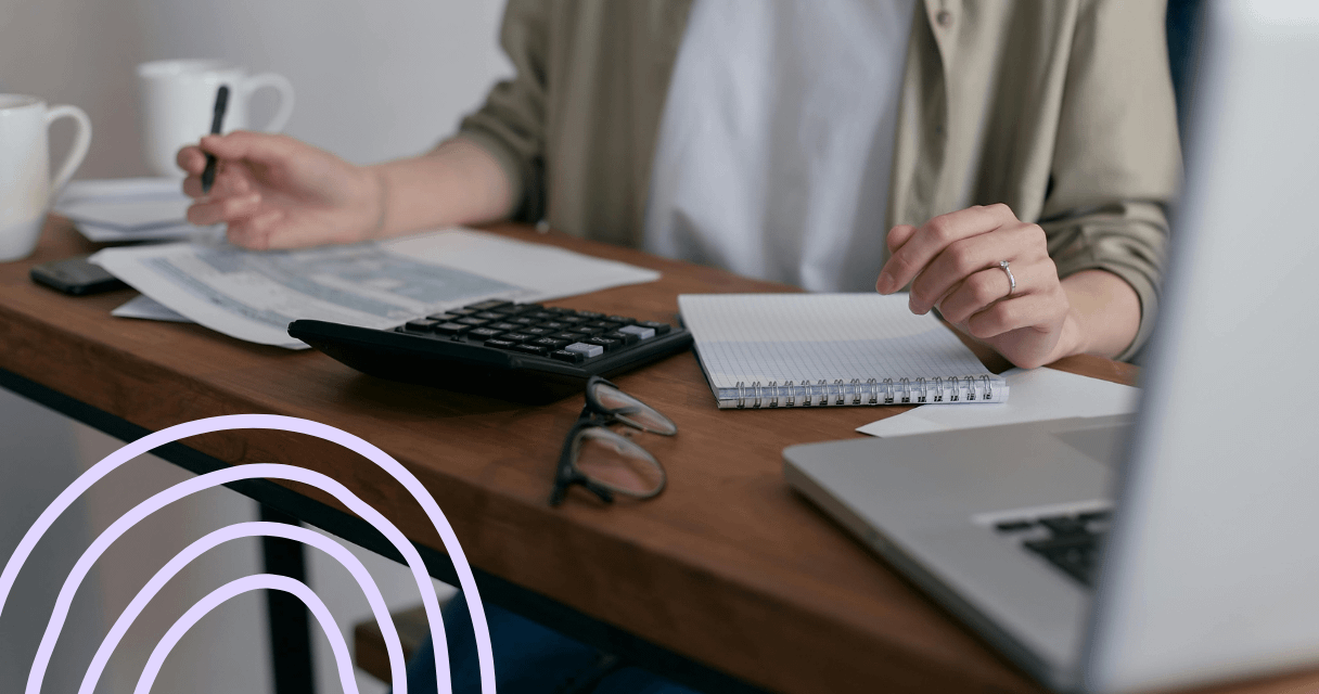 Close-up of a person working at a desk with a calculator, notebook, and documents, emphasizing financial planning and budgeting. A laptop and a pair of glasses are also visible on the wooden surface